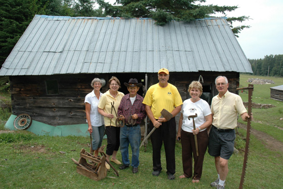 Group in front of small log building, holding tools made by hand in the blacksmith shop