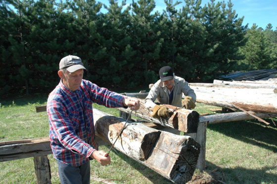 Two men scraping dovetailed round logs with scrapers